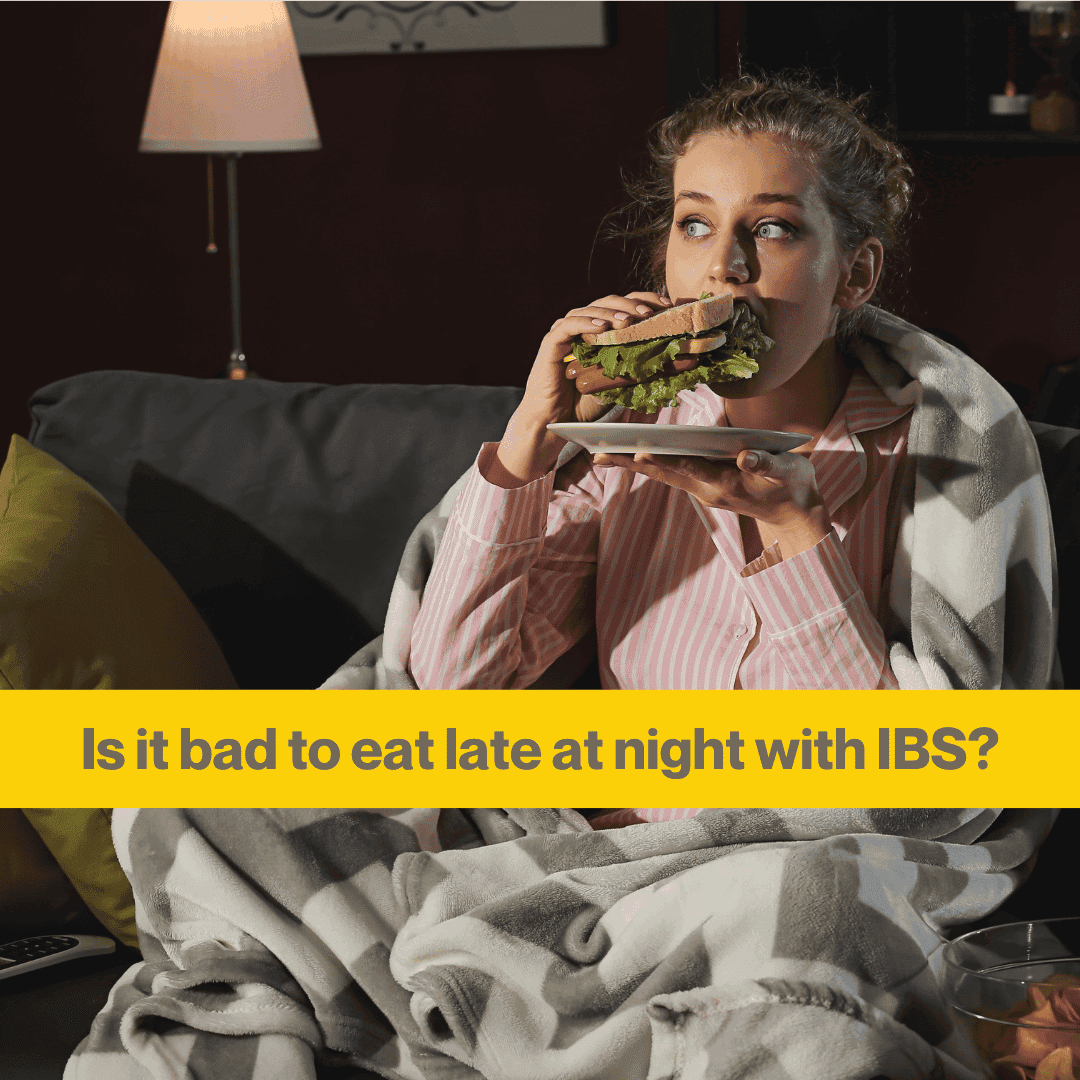 A young white woman sits on a sofa in a darkened room wearing pajamas and wrapped in a blanket. she's eating a sandwich, looking to the side. Words - is it bad to eat late at night with IBS