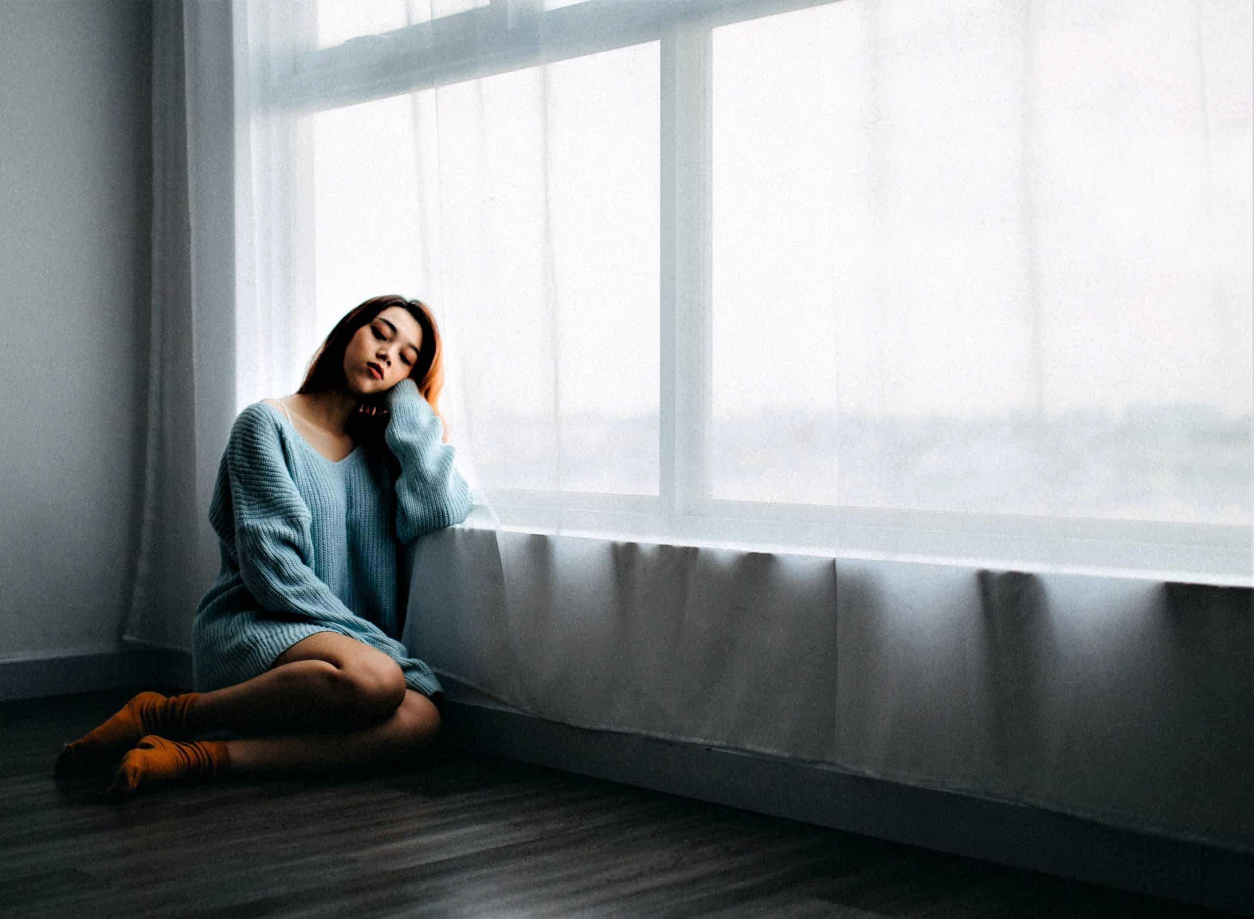 woman sitting on the floor looking sad in an empty room next to big window and a white curtain