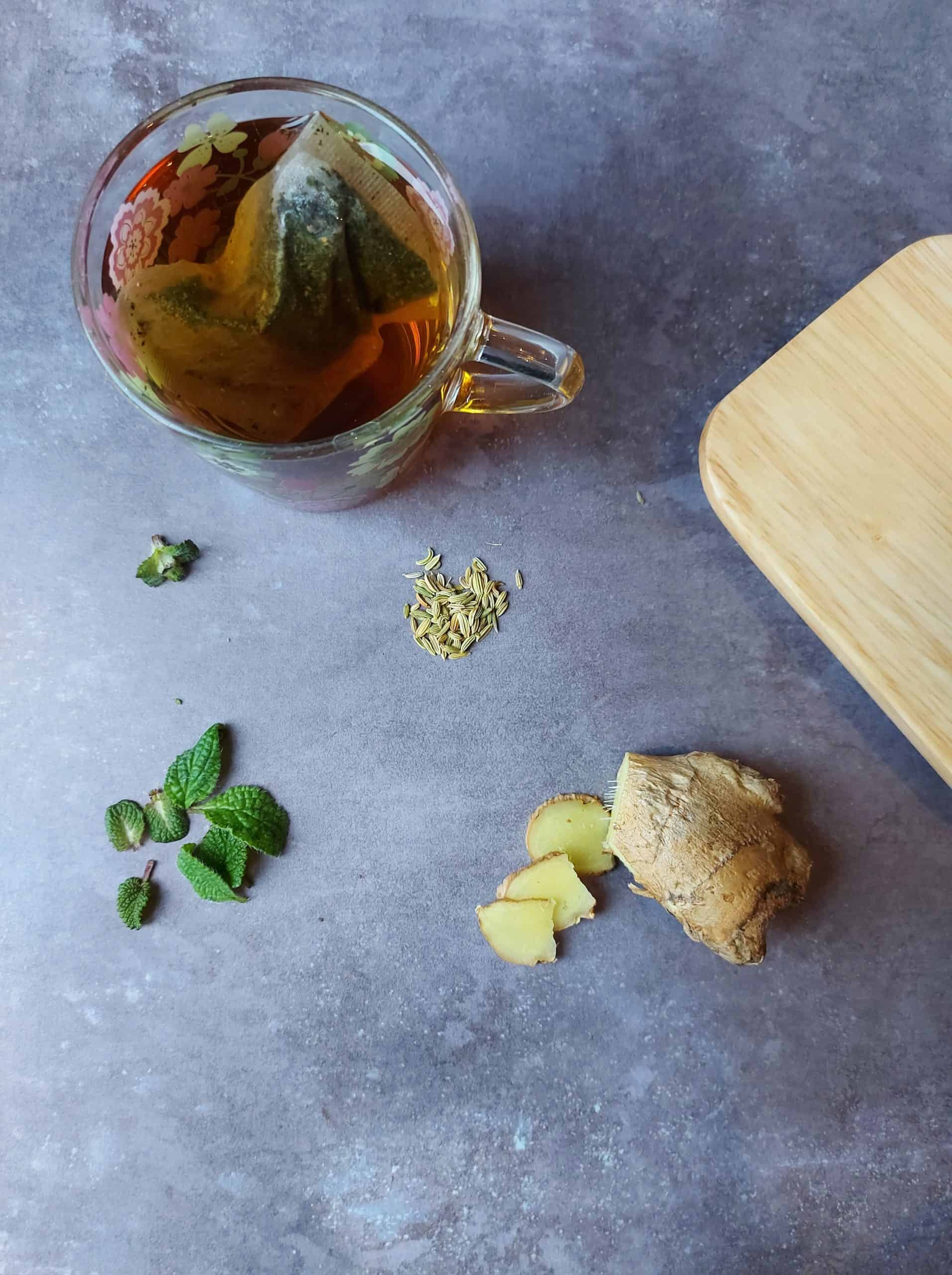 cut ginger root, some lemon balm leaves, small pile of fennel seeds, and a herb tea in a glass, against a dark grey background