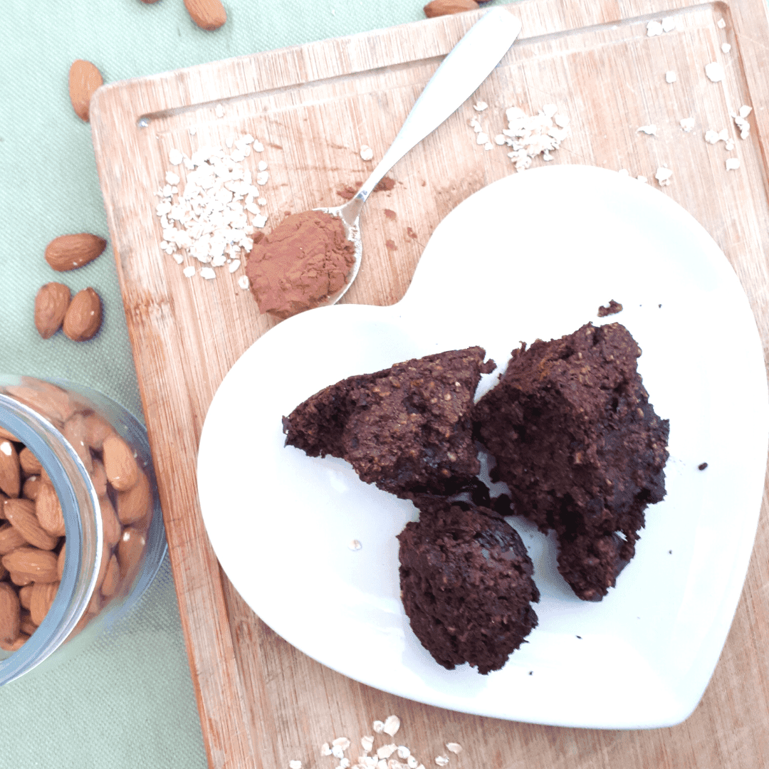 chocolate brownie broken into pieces on a white heart shaped place. Plate is on a wooden board with some oats, next to almonds in a glass jar, and a teaspoon of cocoa powder.