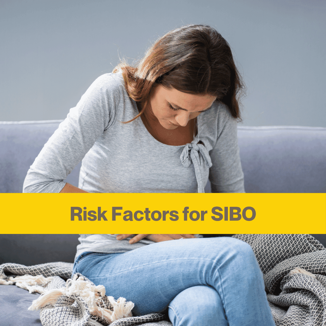 white woman with mid length brown hair wearing light blue jeans and a light grey long sleeved t shirt sitting on grey sofa holding her stomach and looking down.